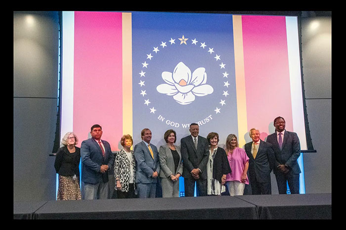 The flag commission with MDAH Director Katie Blount standing in front of the In God We Trust flag projected onto a screen